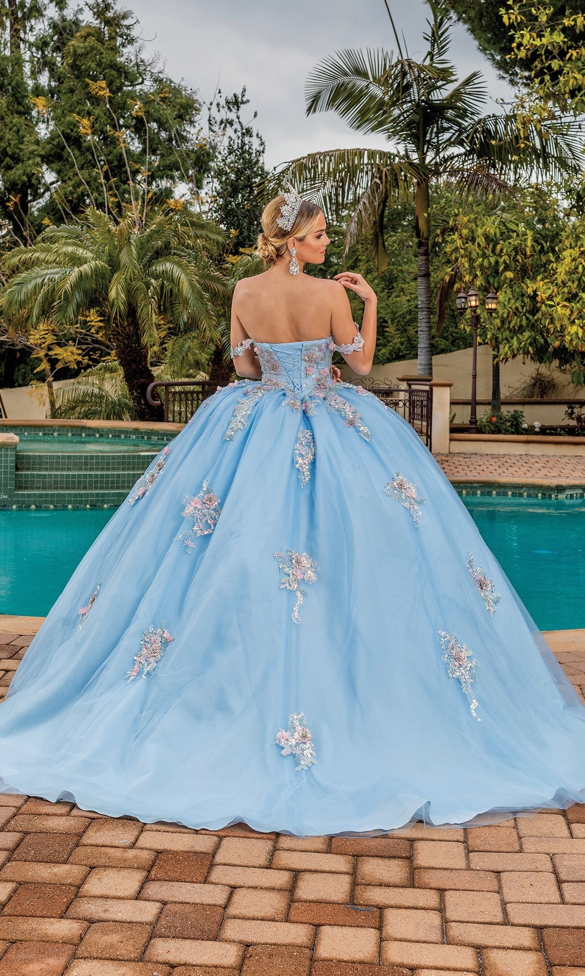 A woman stands by a pool, facing away, wearing the Quinceanera Dress 1845 By Dancing Queen—a light blue off-the-shoulder A-line ball gown with floral embellishments.